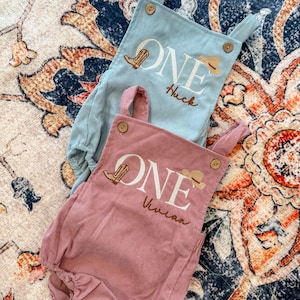 May include: Two corduroy baby rompers, one light blue and one dusty rose. Each features the word "ONE" with a cowboy boot and hat graphic, plus a name. The light blue romper says "Huck", and the dusty rose romper says "Vivian".