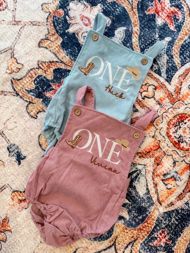 May include: Two corduroy baby rompers, one light blue and one dusty rose. Each features the word "ONE" with a cowboy boot and hat graphic, plus a name. The light blue romper says "Huck", and the dusty rose romper says "Vivian".
