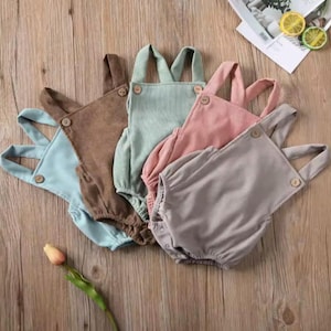May include: Five pastel-coloured corduroy baby rompers in light blue, brown, mint green, pink, and grey. Each has wooden buttons and adjustable straps. The garments are arranged on a wooden surface.