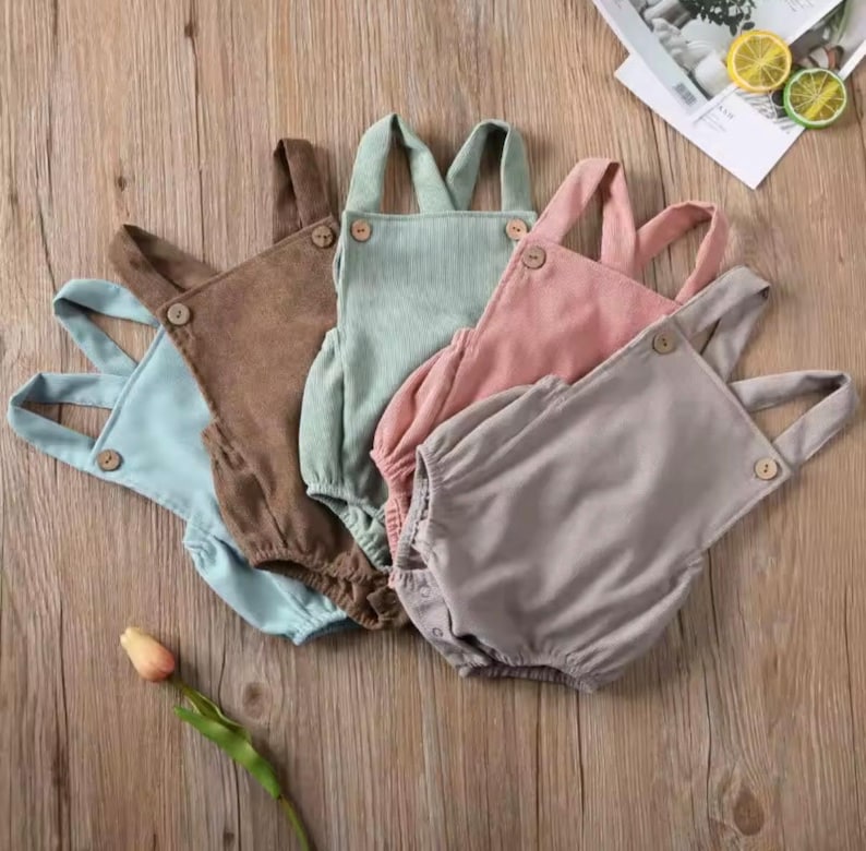 May include: Five pastel-coloured corduroy baby rompers in light blue, brown, mint green, pink, and grey. Each has wooden buttons and adjustable straps. The garments are arranged on a wooden surface.
