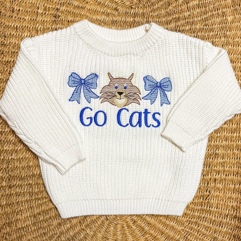 May include: White knitted sweater with a blue embroidered cat and two blue bows. The sweater has the text 'Go Cats' embroidered in blue below the cat.