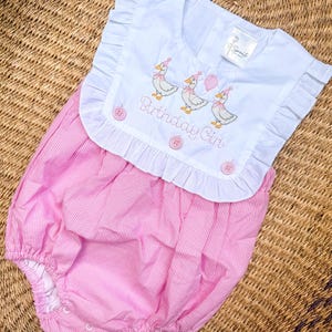 May include: A baby's pink and white gingham romper with a white collar. The collar is embroidered with three ducks wearing party hats and holding balloons, with the text "Birthday Girl". The romper has ruffled edges and pink buttons.