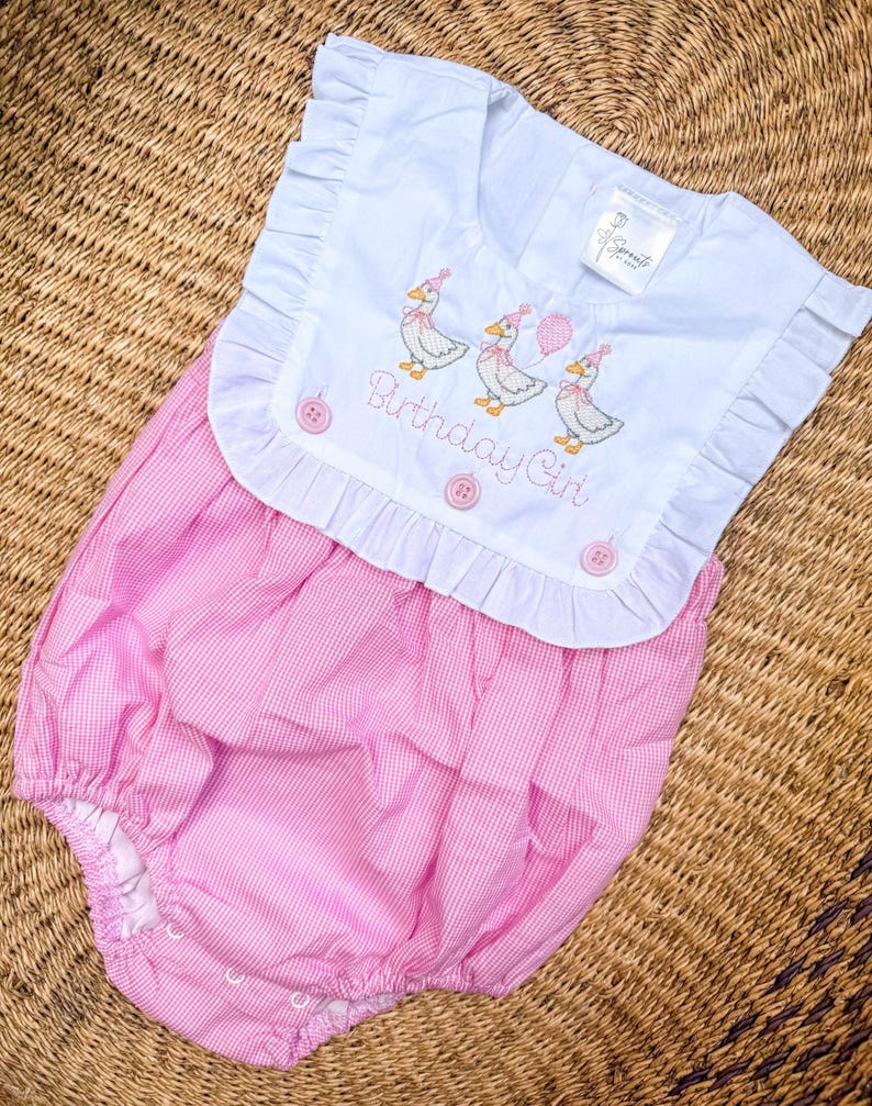 May include: A baby's pink and white gingham romper with a white collar. The collar is embroidered with three ducks wearing party hats and holding balloons, with the text "Birthday Girl". The romper has ruffled edges and pink buttons.