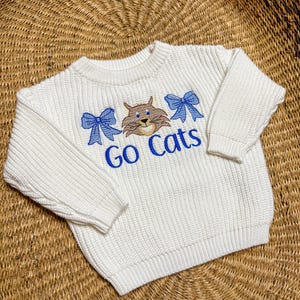 May include: Off-white knit sweater with long sleeves. The sweater features an embroidered design with a brown cat, two blue bows, and the words "Go Cats" in blue. The sweater is made of a textured knit fabric.