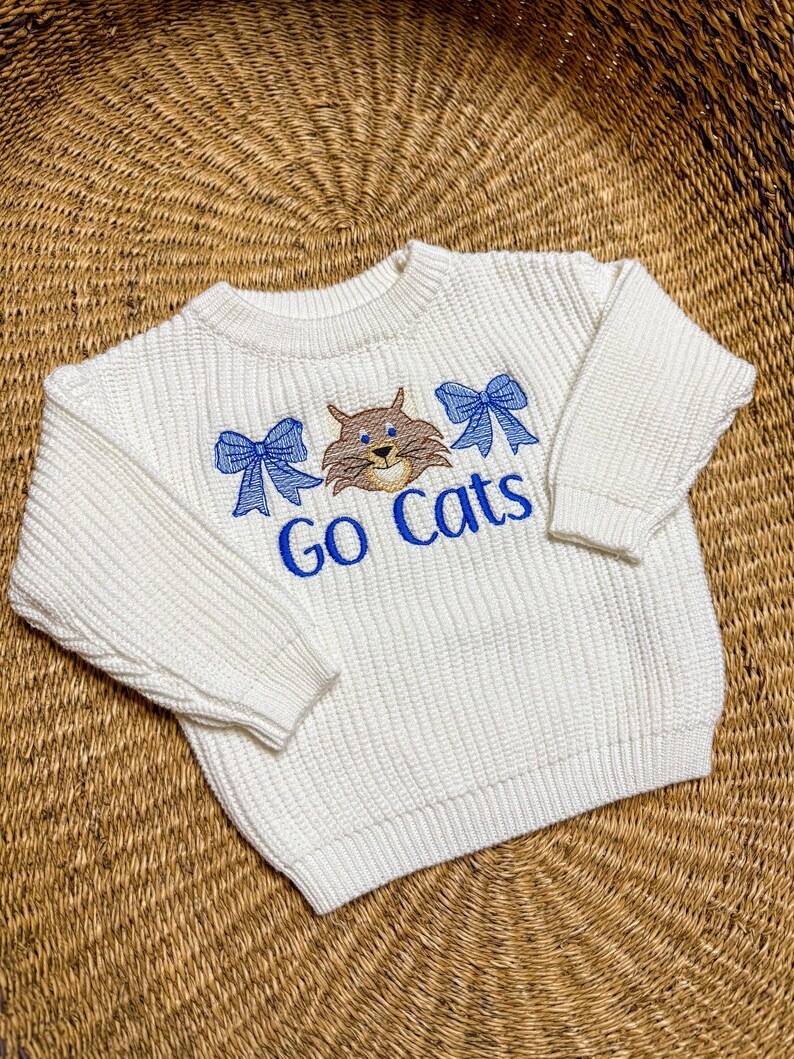 May include: Off-white knit sweater with long sleeves. The sweater features an embroidered design with a brown cat, two blue bows, and the words "Go Cats" in blue. The sweater is made of a textured knit fabric.