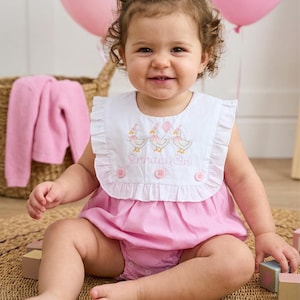 May include: A smiling baby girl wearing a pink gingham romper with a white bib. The bib has embroidered ducks and the words "Birthday Girl." Pink balloons with the number "1" are in the background. A pink sweater and wooden blocks are also visible.