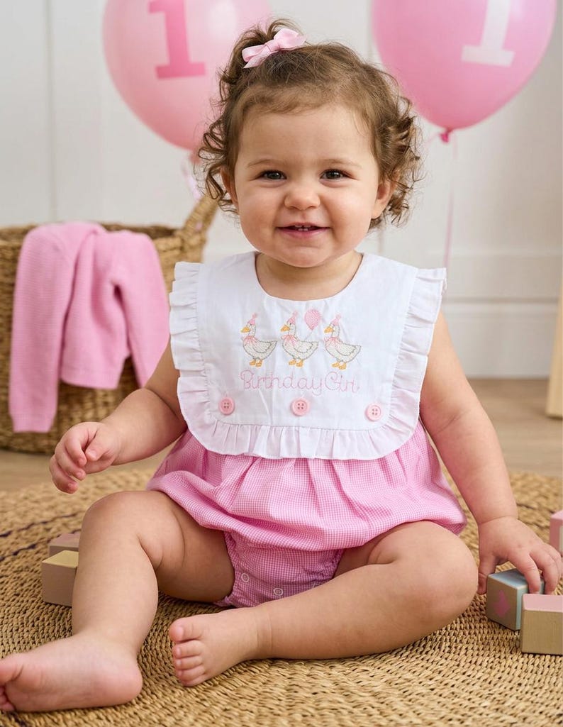 May include: A smiling baby girl wearing a pink gingham romper with a white bib. The bib has embroidered ducks and the words "Birthday Girl." Pink balloons with the number "1" are in the background. A pink sweater and wooden blocks are also visible.