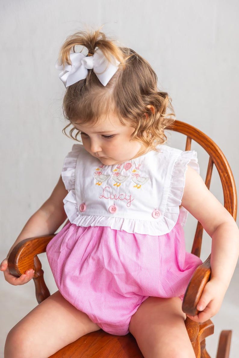 May include: A pink and white gingham romper with a white embroidered bib featuring ducks and the name "Lucy". The romper has ruffled sleeves and is fastened with pink buttons. A white bow adorns the child's hair.