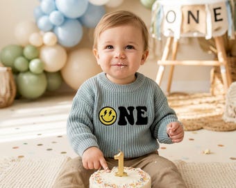May include: A baby wearing a light blue sweater with the word "ONE" and a smiley face graphic, sitting in front of a cake with a number one candle. Balloons in shades of blue and white are in the background.