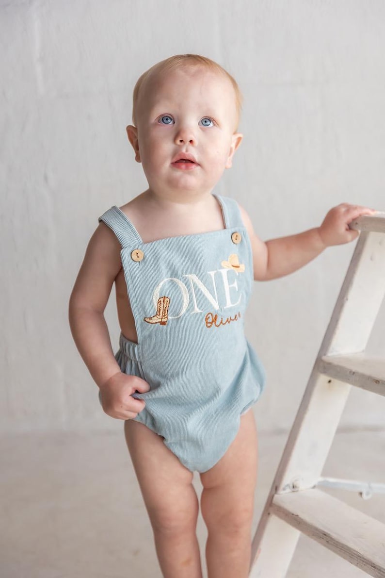 May include: A light blue corduroy romper with the word "ONE" embroidered on the front, along with a cowboy boot and hat. The romper has wooden button closures and is designed for a baby's first birthday. The name "Oliver" is also embroidered.