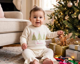 May include: A baby wearing a cream-colored knit sweater and pants set with green Christmas tree embroidery and the name "Josie" in pink script. The baby is sitting near a decorated Christmas tree and wrapped gifts.