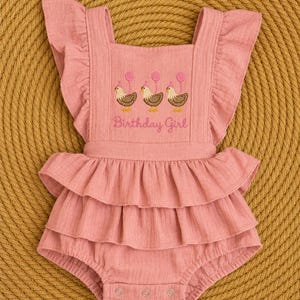 May include: A dusty rose-colored romper featuring ruffled shoulder straps and a double-layered ruffled waist. The chest is embroidered with three brown chickens wearing pink party hats and balloons, with the text Birthday Girl below them.