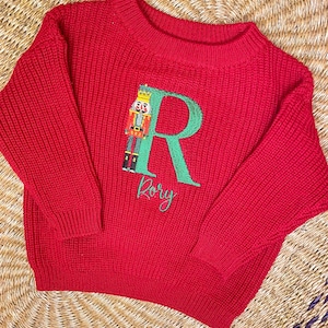 Puede incluir: Un suéter rojo tejido con cuello redondo. El suéter presenta una letra "R" verde bordada con un diseño de cascanueces y el nombre "Rory" en escritura verde. El suéter está hecho de un tejido grueso y texturizado.