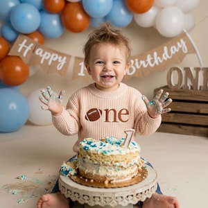 May include: A smiling child wearing a light pink sweater with a football graphic and the word "one". The child is sitting in front of a cake with blue and white frosting. Balloons and a banner that says "Happy 1st Birthday" are in the background.