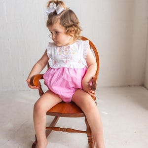 May include: A little girl wearing a white top with pink shorts, sitting in a wooden rocking chair. The top has a ruffle collar and embroidery with the name "Lucy". The shorts are a bright pink color.