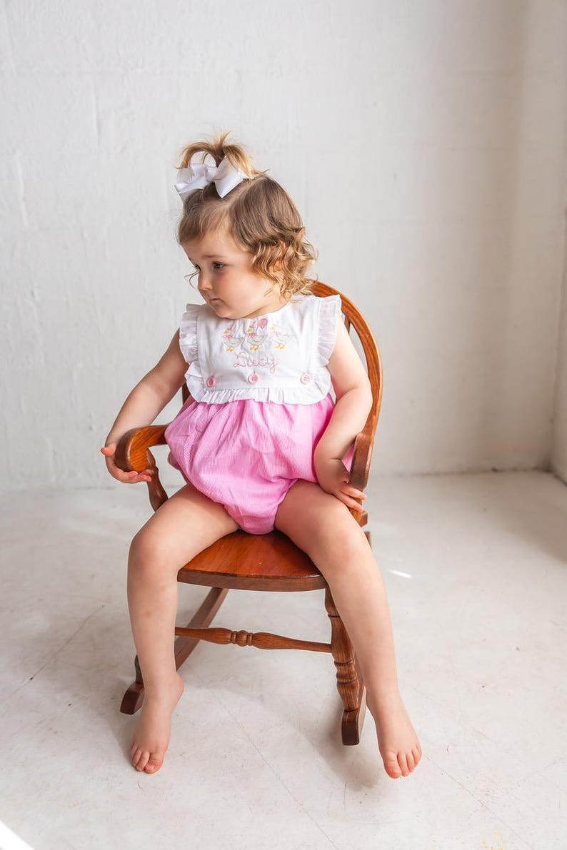 May include: A little girl wearing a white top with pink shorts, sitting in a wooden rocking chair. The top has a ruffle collar and embroidery with the name "Lucy". The shorts are a bright pink color.