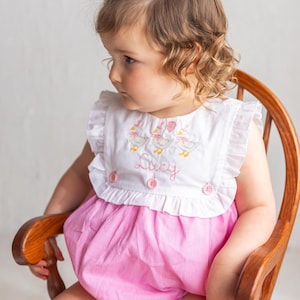May include: A baby girl in a pink and white gingham romper with a white bib top. The bib is embroidered with ducks and the name "Lucy". She wears a white bow and sits in a wooden rocking chair.