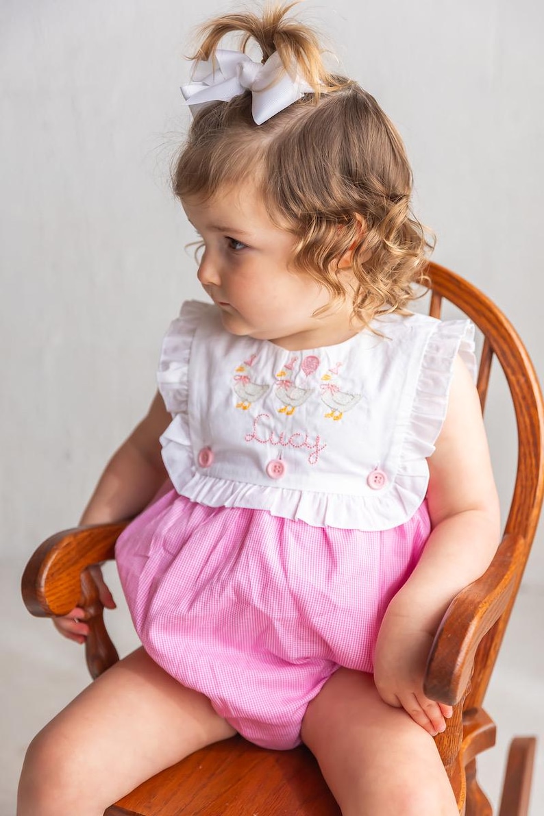 May include: A baby girl in a pink and white gingham romper with a white bib top. The bib is embroidered with ducks and the name "Lucy". She wears a white bow and sits in a wooden rocking chair.
