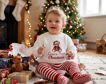 Op de afbeelding: Een baby draagt een witte trui met een teddybeer en de tekst "Liam's First Christmas" geborduurd, gecombineerd met rood-wit gestreepte broek. De baby is omringd door ingepakte cadeaus en een kerstboom.