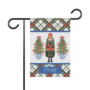 May include: A garden flag featuring a watercolor design of a nutcracker soldier, two Christmas trees, and the name "Crosby" on a blue banner. The background has a green and red plaid pattern.