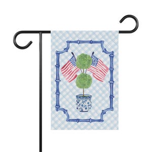 May include: A garden flag featuring a watercolor design. The flag displays a blue bamboo-style frame with a light blue gingham background. Inside the frame are two American flags and a topiary in a blue and white pot.