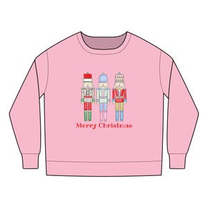 Puede incluir: Sudadera rosa claro con tres soldados cascanueces ilustrados en rojo, azul y verde. Las palabras "Merry Christmas" están impresas en rojo debajo. La sudadera tiene mangas largas y cuello redondo.