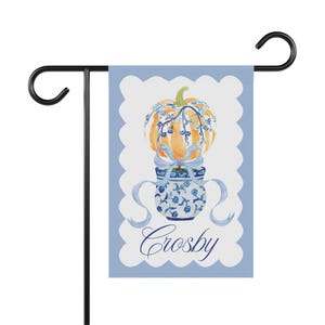 May include: A garden flag with a watercolour design of an orange pumpkin with blue floral accents and a blue ribbon, atop a blue and white vase. The flag has a light blue scalloped border and the name "Crosby" in script. The flag is mounted on a black metal stand.