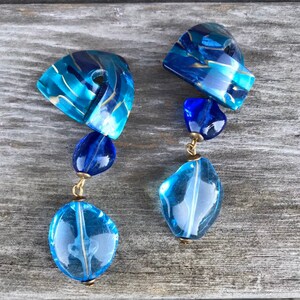 May include: A pair of blue and gold dangle earrings. Each earring features a large, irregular-shaped, light blue bead at the bottom, connected by gold-colored metal to smaller blue beads and a geometric top piece with blue and gold swirls.