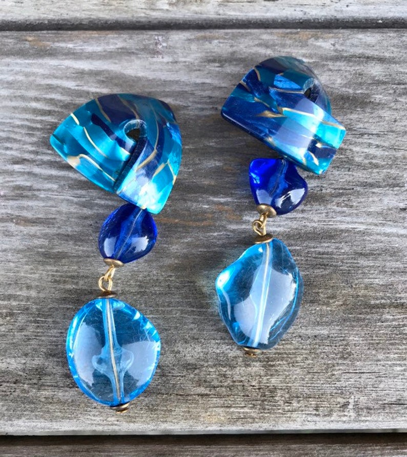 May include: A pair of blue and gold dangle earrings. Each earring features a large, irregular-shaped, light blue bead at the bottom, connected by gold-colored metal to smaller blue beads and a geometric top piece with blue and gold swirls.
