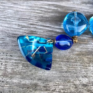 May include: A pair of blue earrings with a geometric design. The earrings feature a combination of translucent and opaque blue elements, connected by gold-colored metal. The design includes a large, angular pendant and smaller, round beads.