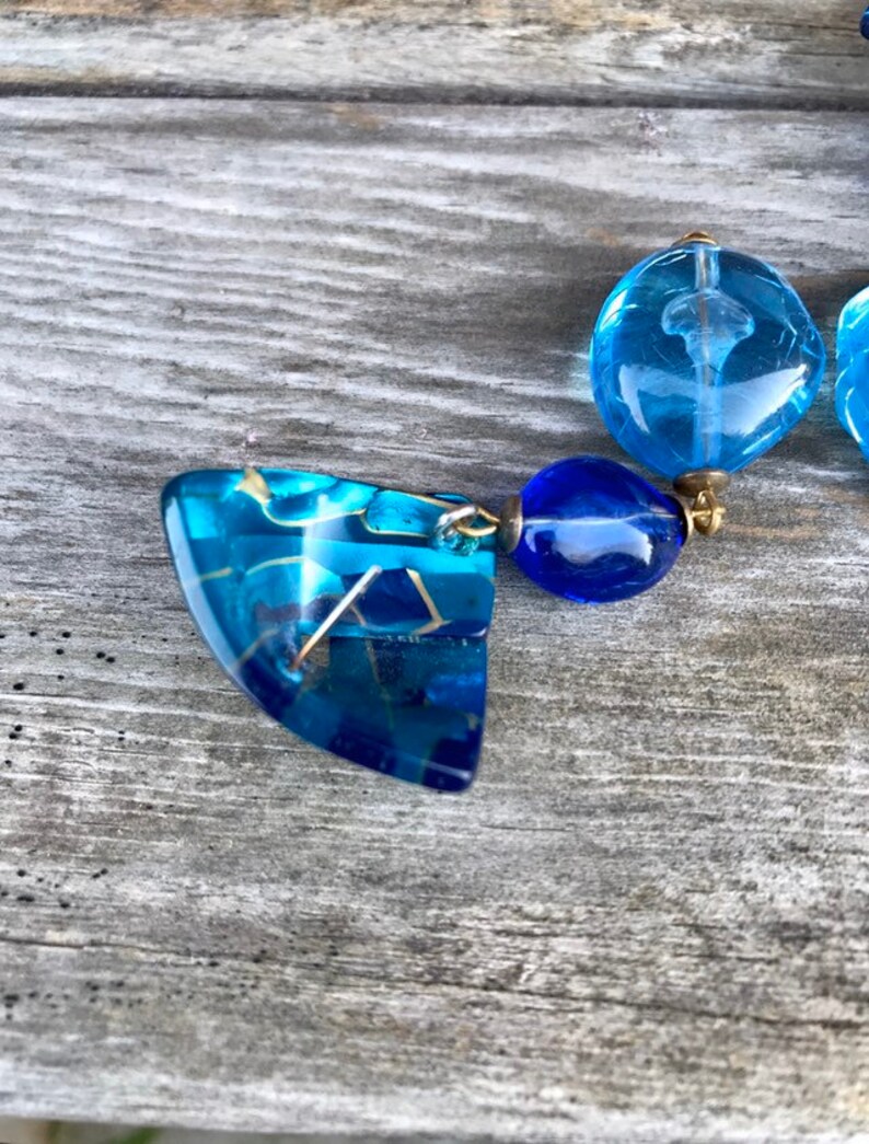 May include: A pair of blue earrings with a geometric design. The earrings feature a combination of translucent and opaque blue elements, connected by gold-colored metal. The design includes a large, angular pendant and smaller, round beads.