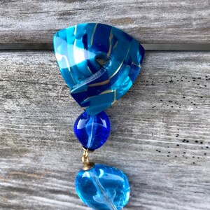 May include: A vintage blue and gold-toned pendant. The top section is a triangular shape with a marbled effect. Two blue glass beads hang below, connected by gold-colored metal. The bottom bead is a lighter shade of blue.