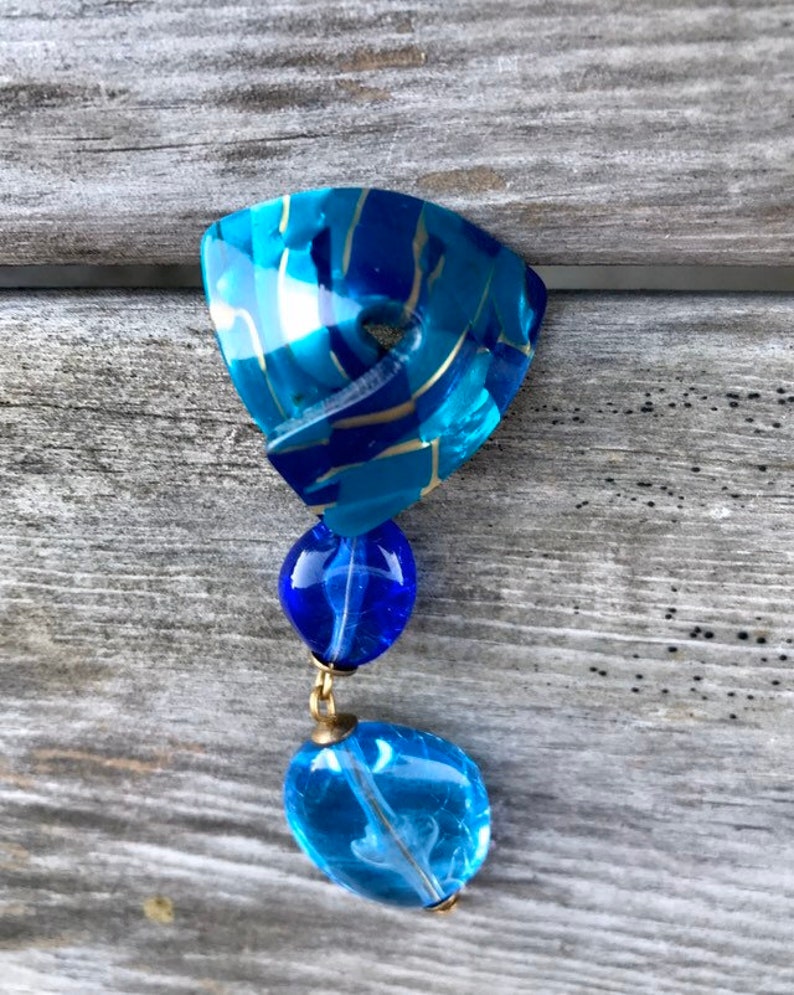 May include: A vintage blue and gold-toned pendant. The top section is a triangular shape with a marbled effect. Two blue glass beads hang below, connected by gold-colored metal. The bottom bead is a lighter shade of blue.