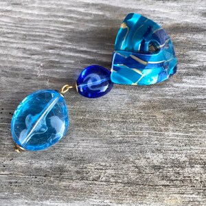 May include: A unique, handcrafted earring featuring three blue, translucent, and gold-accented beads. The largest bead is an oval shape with a white swirl design. The other two beads are round and connected with gold-colored wire.