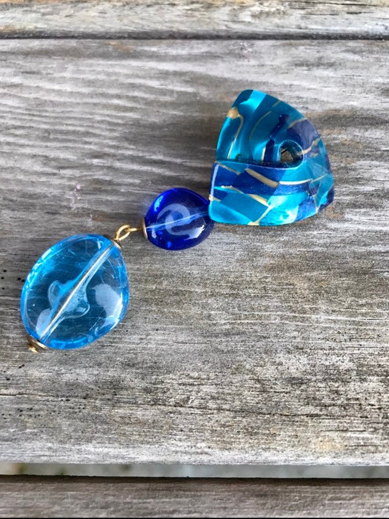 May include: A unique, handcrafted earring featuring three blue, translucent, and gold-accented beads. The largest bead is an oval shape with a white swirl design. The other two beads are round and connected with gold-colored wire.