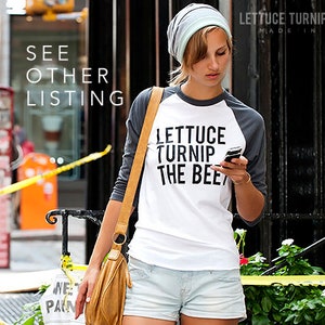 May include: A woman wearing a white and gray baseball tee shirt with the text "Lettuce Turnip The Beet" printed on it. She is wearing a light blue beanie and denim shorts. She is holding a black cell phone in her right hand.