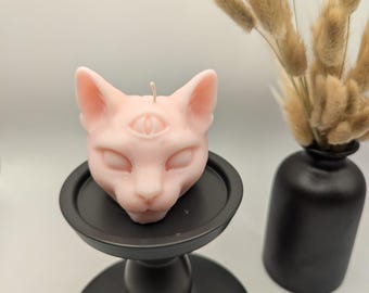 Puede incluir: Una vela de cabeza de gato rosa con un tercer ojo en la frente, sentada en un candelero negro.