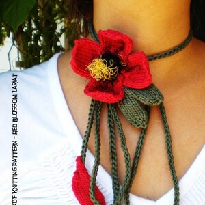 Pode incluir: Uma flor vermelha de tricô com folhas verdes, presa a um cordão de tricô verde, usada como colar. O colar se chama "Red Blossom Lariat" e está disponível como um padrão de tricô PDF.
