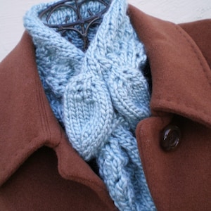 May include: A light blue knitted scarf with a cable stitch pattern. The scarf is draped over a brown coat.