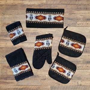 May include: A kitchen set consisting of a rectangular towel, a square towel, an oven mitt, and two square pot holders. Each item is black with a horizontal band featuring a geometric pattern in orange, brown, and white with dreamcatcher icons.