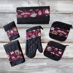 May include: Set of black and maroon kitchen accessories with a Montana Grizzlies theme. The set includes a potholder, oven mitt, and two dish towels. The potholders and oven mitt feature a quilted design.