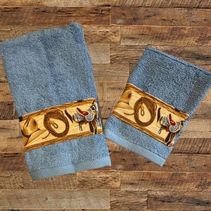 May include: Two blue hand towels with a brown, yellow and blue western-themed border. The border features a cowboy hat, rope and a saddle.