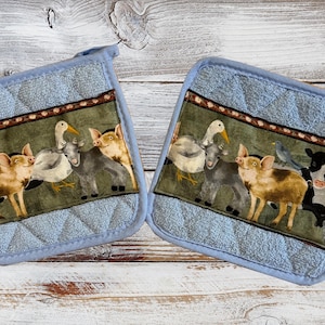 May include: Two blue quilted pot holders with a farm animal design. The design features a goose, pig, donkey, and cow. The pot holders have a brown and white trim.