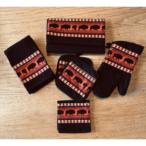 May include: A kitchen set featuring a black theme with a decorative band of bison silhouettes on an orange background. The set includes a towel, oven mitt, pot holders, and a folded item, likely a dish towel or pot holder.