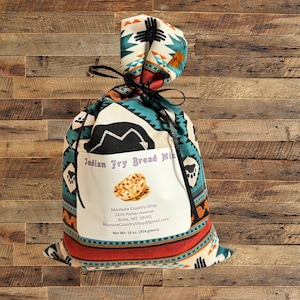 May include: A white bag with a colourful Native American pattern, tied with a black ribbon. The bag contains Indian Fry Bread Mix and has a label with the name of the product, the shop's name, address, email, and net weight: 454 grams (16 oz).