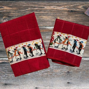 May include: Two red kitchen towels with a white border and a pattern of penguins wearing Santa hats. The penguins are holding hands and walking in a line.