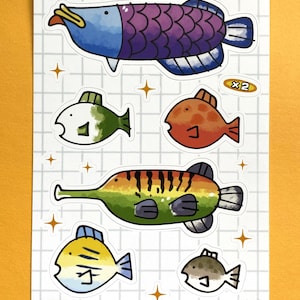 Puede incluir: Un conjunto de seis pegatinas de peces coloridos con fondo blanco y un patrón de cuadrícula. Las pegatinas presentan una variedad de peces, incluyendo un pez largo y rayado, un pez redondo y naranja, y un pez azul y morado con un cuerpo largo y delgado. Las pegatinas también incluyen una pequeña estrella blanca y el texto "x2" en un círculo negro.