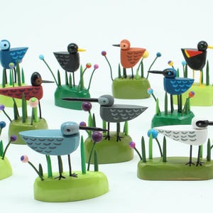 Puede incluir: Una colección de 12 figuritas de pájaros de madera pintadas a mano. Cada pájaro está posado sobre una base verde con hierba verde y flores de colores. Los pájaros están pintados en varios colores, incluyendo azul, rojo, amarillo, negro y blanco.