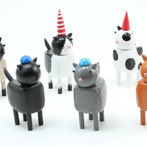 Op de afbeelding: Een verzameling grillige houten dierenfiguren, elk geschilderd in een uniek kleurenschema. De figuren omvatten schapen, katten en koeien, allemaal met feestelijke hoeden. Het kleurenpalet omvat zwart, wit, grijs en oranje, met accenten van rood en blauw.