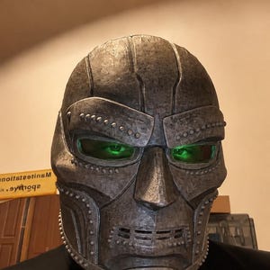 May include: A metallic, weathered-looking full-face mask with a textured, dark grey finish. The mask features prominent rivet details along the brow and jawline, a central nose piece, and horizontal vents at the mouth. Bright green lenses cover the eyes.